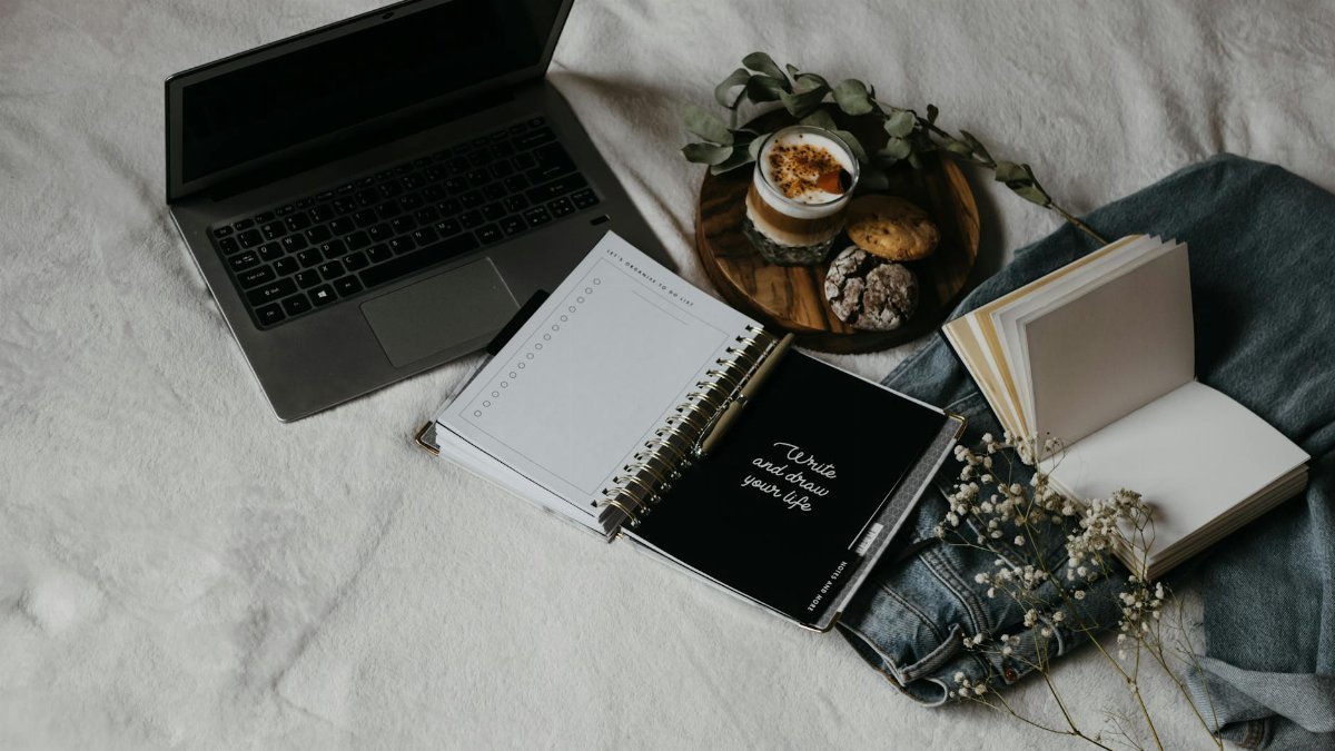 A cozy flat lay of a laptop, journals, and snacks on a bed for a relaxed work-from-home setup.