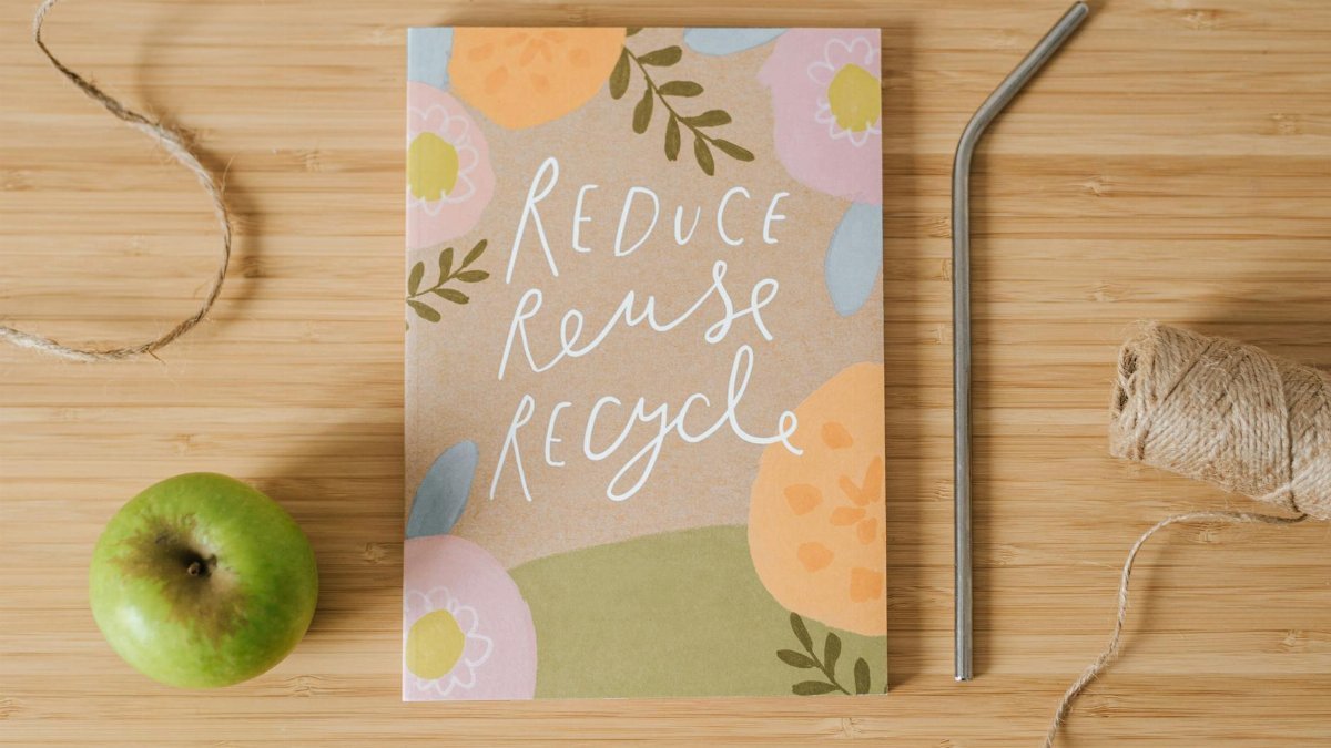 Top view of book with Reduce Reuse Recycle inscriptions near green apple and bobbin of thread on table