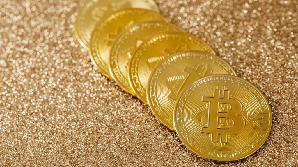A stack of gold Bitcoin coins placed on a shimmering gold glitter background.