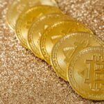 A stack of gold Bitcoin coins placed on a shimmering gold glitter background.