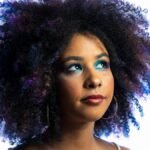 Close-up portrait of a woman with vibrant afro hair and striking blue eye makeup on a white background.