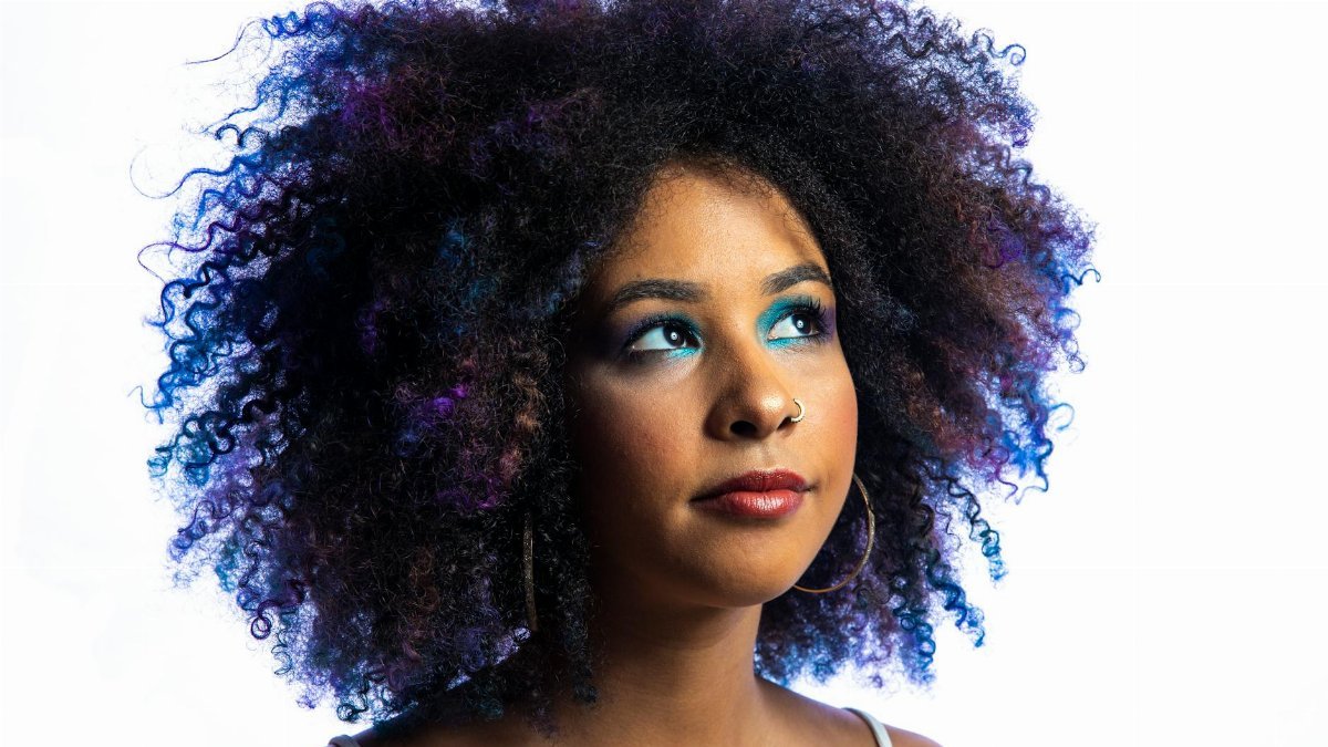 Close-up portrait of a woman with vibrant afro hair and striking blue eye makeup on a white background.