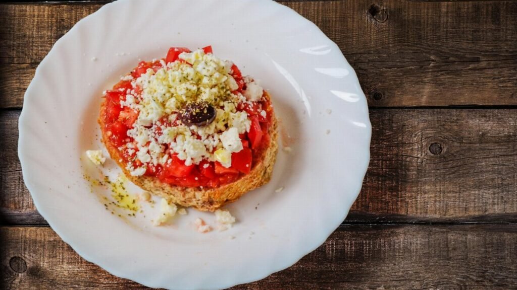 A close-up of a classic Greek Dakos salad with feta, tomato, and olive on a wooden table.