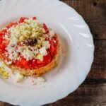 A close-up of a classic Greek Dakos salad with feta, tomato, and olive on a wooden table.