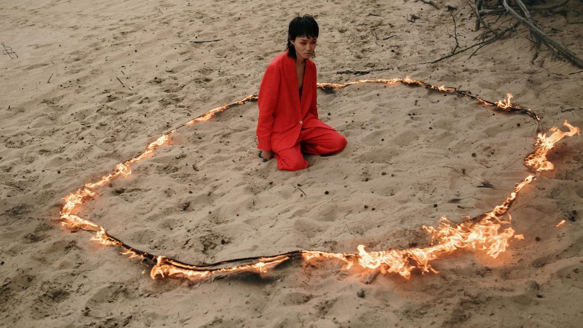 Individual in red suit sits in a contemplative pose, surrounded by fire on a sandy beach.
