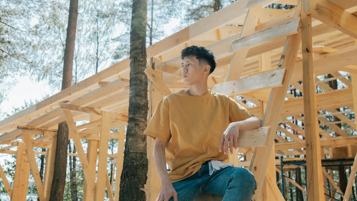 A young man sitting on a ladder at a forest construction site surrounded by wooden beams.