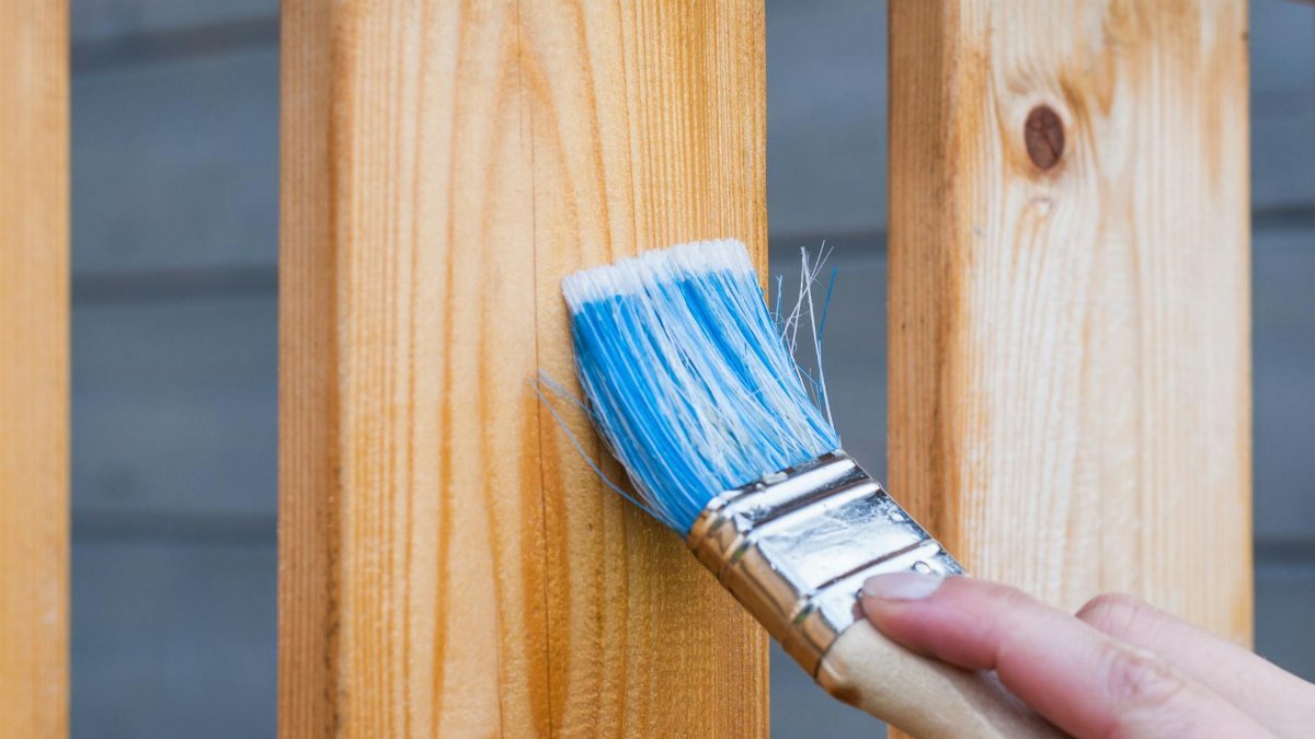 A hand painting a wooden fence plank with a blue paintbrush outdoors. Perfect for DIY and renovation themes.