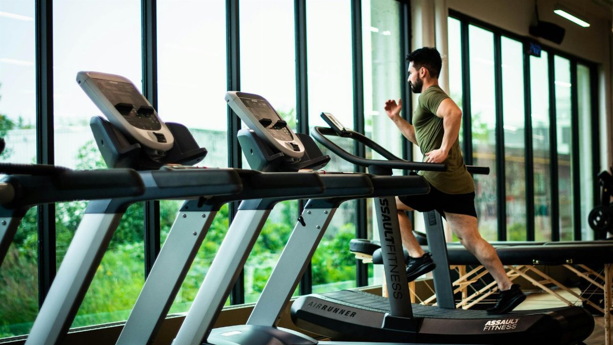 Young man workouts on treadmill in modern gym with large windows and natural light.