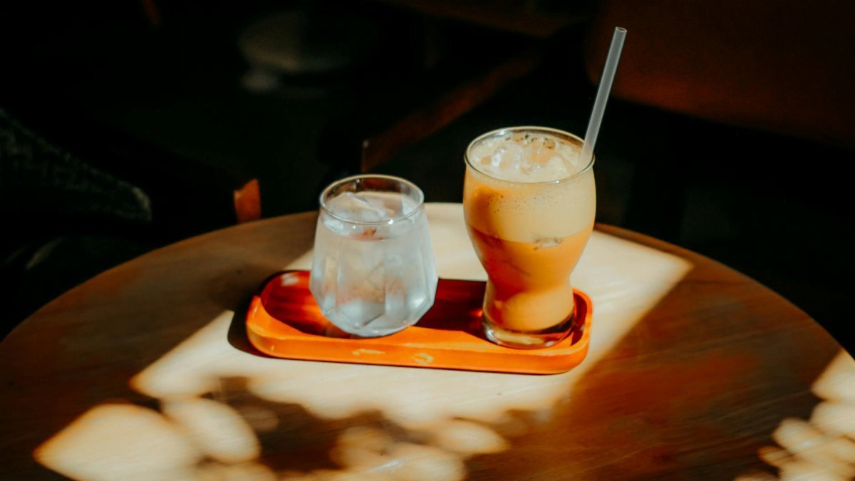 A refreshing iced coffee and glass of water sit on a sunlit wooden table indoors.