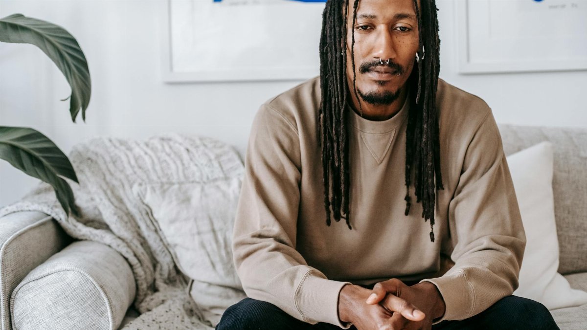 Crop thoughtful African American male in casual sweater with dreadlocks sitting with hands clasped on comfy couch in modern living room