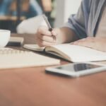 A woman writes in a notebook at a café table with a coffee and smartphone nearby.