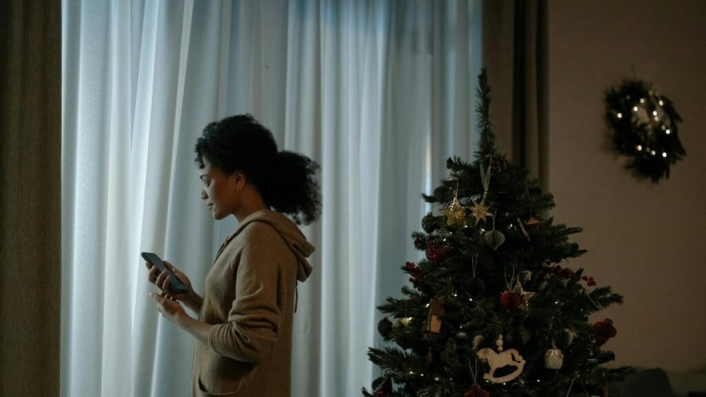Young woman looks outside while holding a smartphone near a decorated Christmas tree indoors.