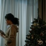 Young woman looks outside while holding a smartphone near a decorated Christmas tree indoors.