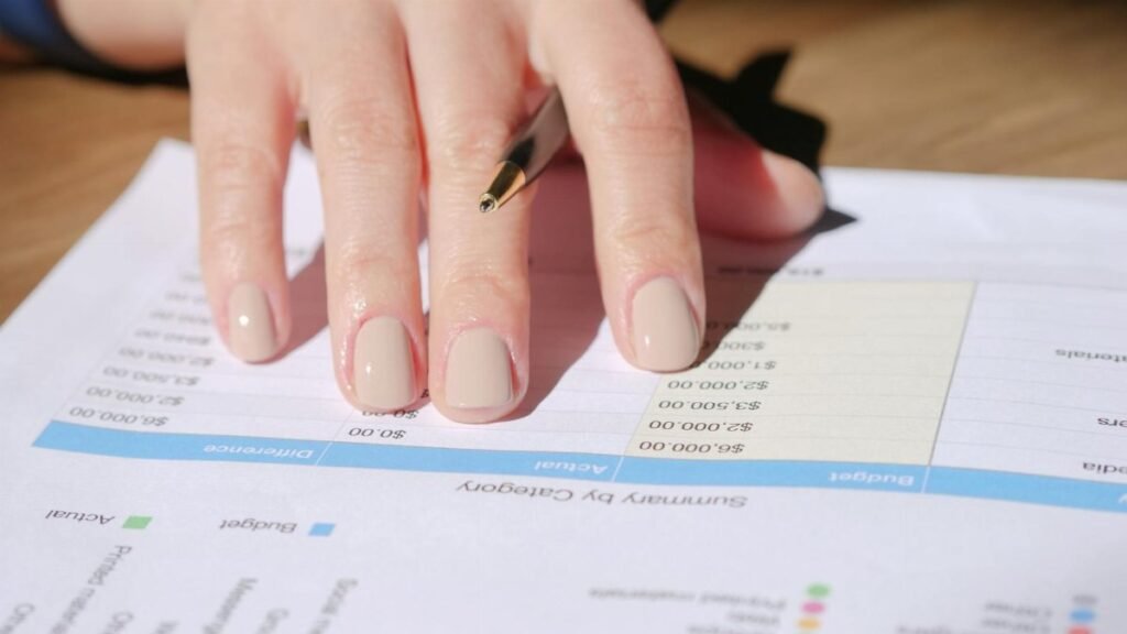 A hand writing on a financial document with a pen in a sunlit office space, showing graphs and data.