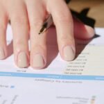 A hand writing on a financial document with a pen in a sunlit office space, showing graphs and data.