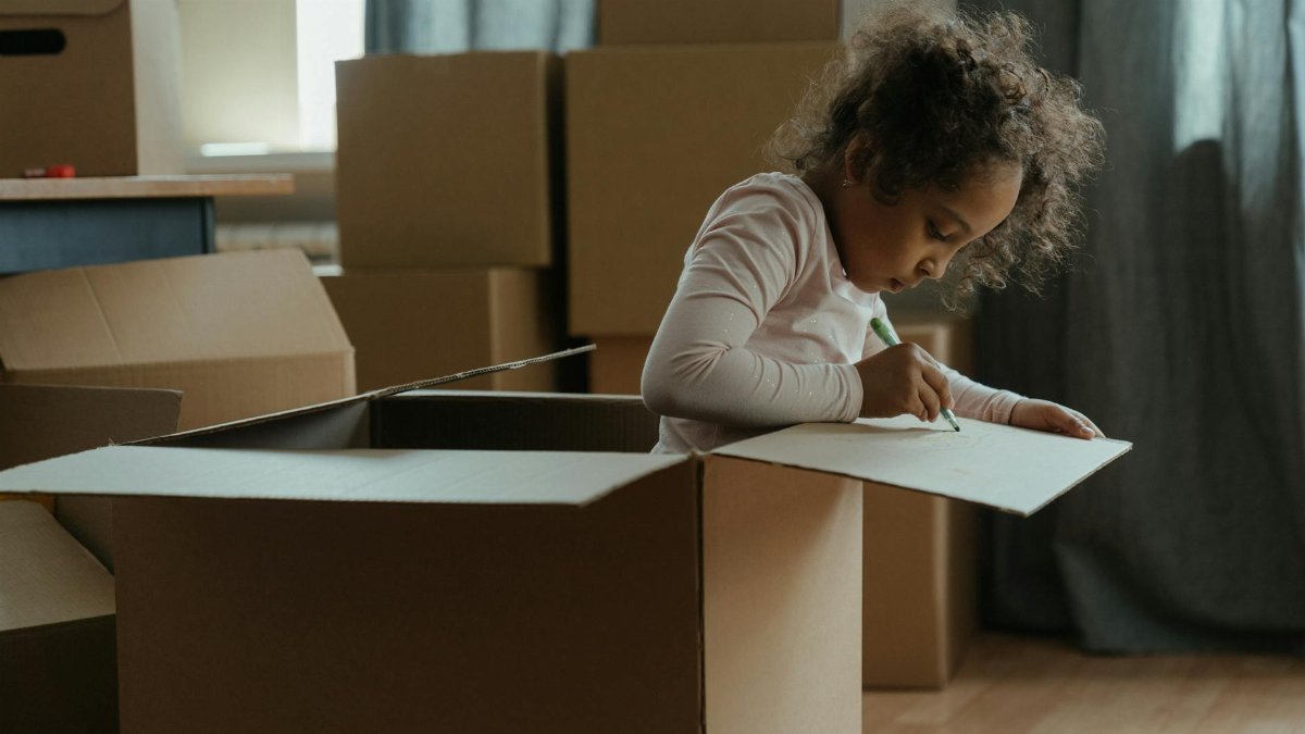 A young child creatively drawing while sitting in a cardboard box surrounded by moving boxes indoors.