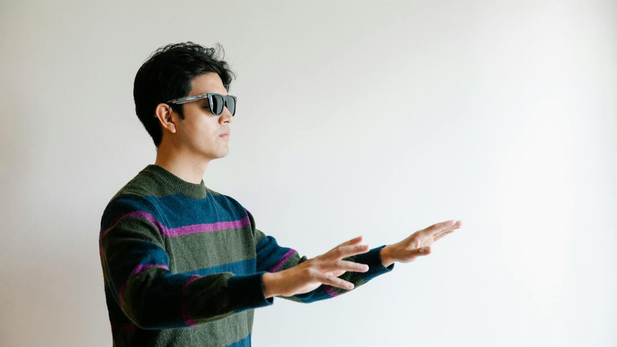 A visually impaired man using his hands to navigate an indoor space, wearing sunglasses.