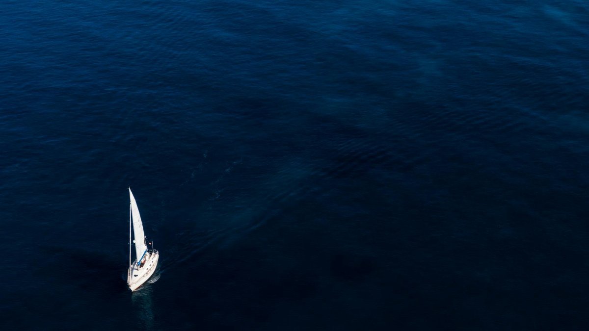 A lone sailboat glides on the vast, blue ocean under clear skies.