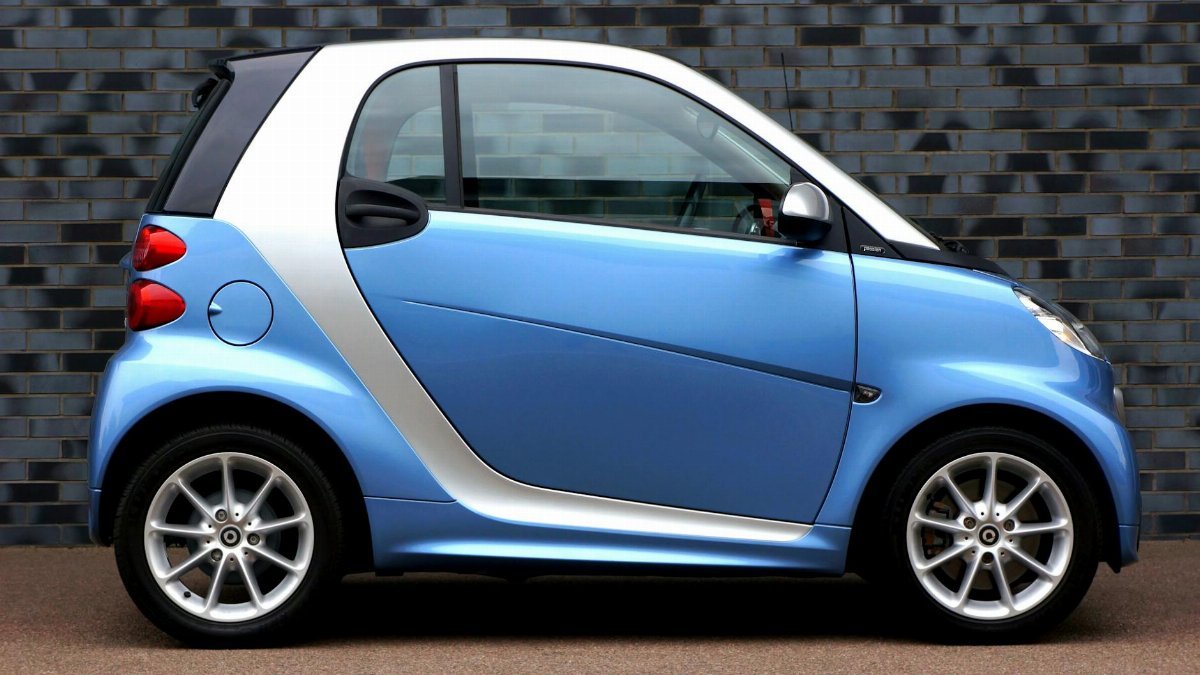 Side view of a modern blue smart car against a brick wall, showcasing compact design.
