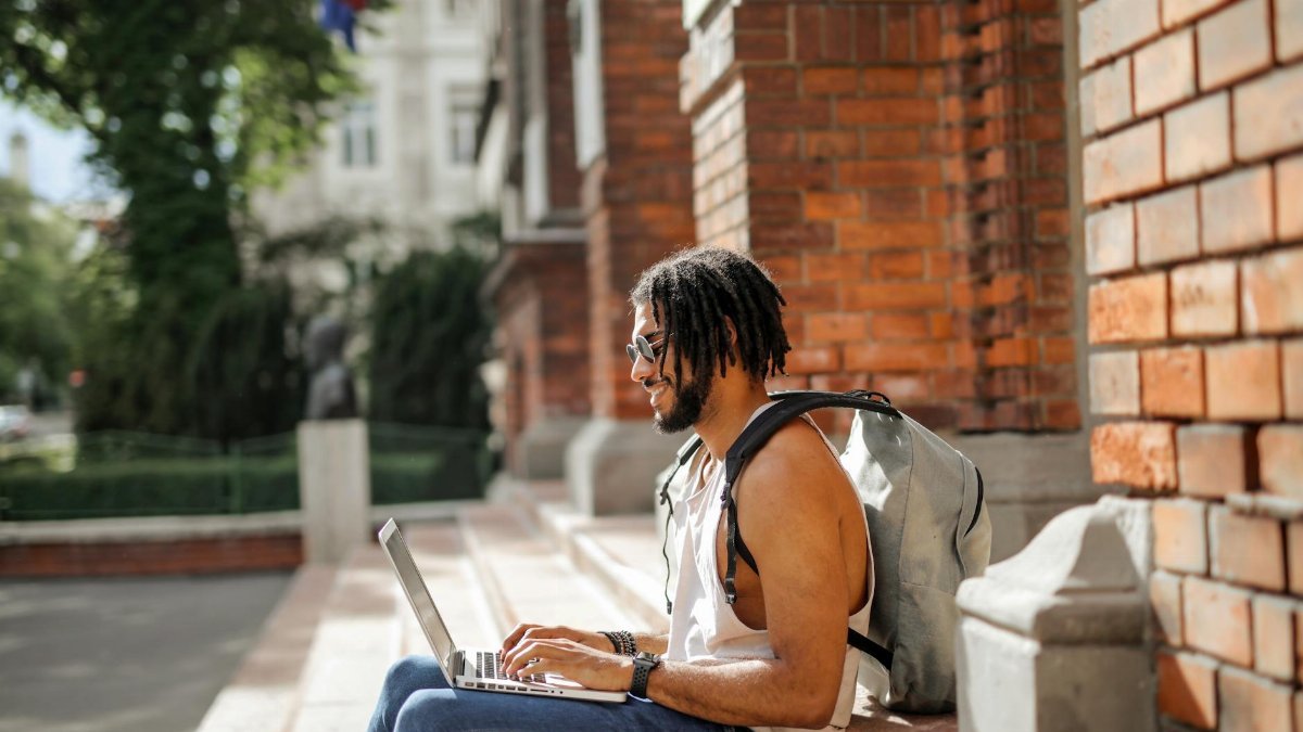 A young man in casual attire sits on outdoor steps working on his laptop with urban background.