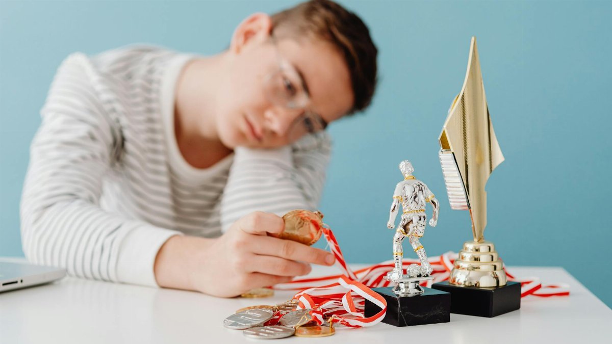 A thoughtful teenager sits by a table with various trophies and medals.