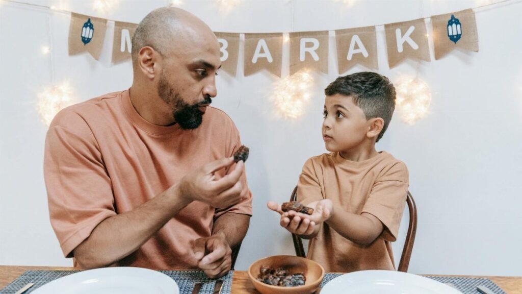 A father and son share dates during an Eid Mubarak celebration indoors.