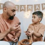 A father and son share dates during an Eid Mubarak celebration indoors.
