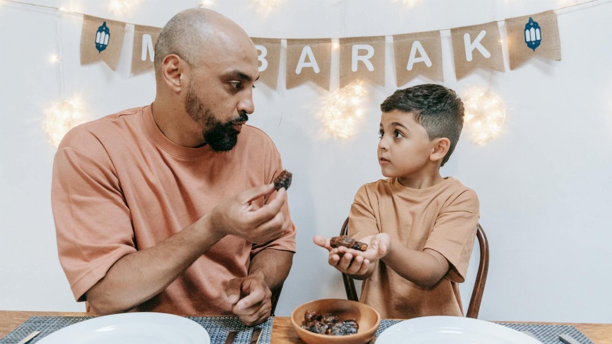 A father and son share dates during an Eid Mubarak celebration indoors.
