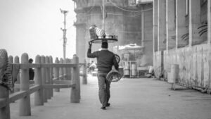 A street vendor walks with goods on his head along the ghats in Varanasi, capturing a timeless black and white scene.