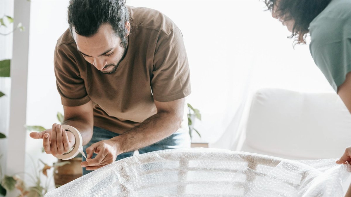 A couple wrapping furniture with bubble wrap and adhesive tape in a bright home setting, preparing to move.
