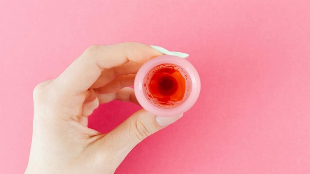A detailed view of a menstrual cup with red liquid held by a hand against a pink backdrop, symbolizing menstruation.