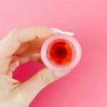 A detailed view of a menstrual cup with red liquid held by a hand against a pink backdrop, symbolizing menstruation.