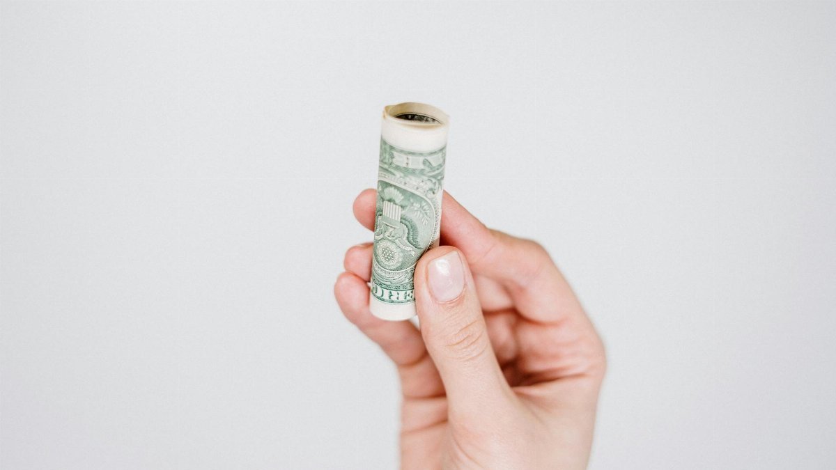 A hand holding a rolled US dollar bill against a plain background, symbolizing finance and savings.
