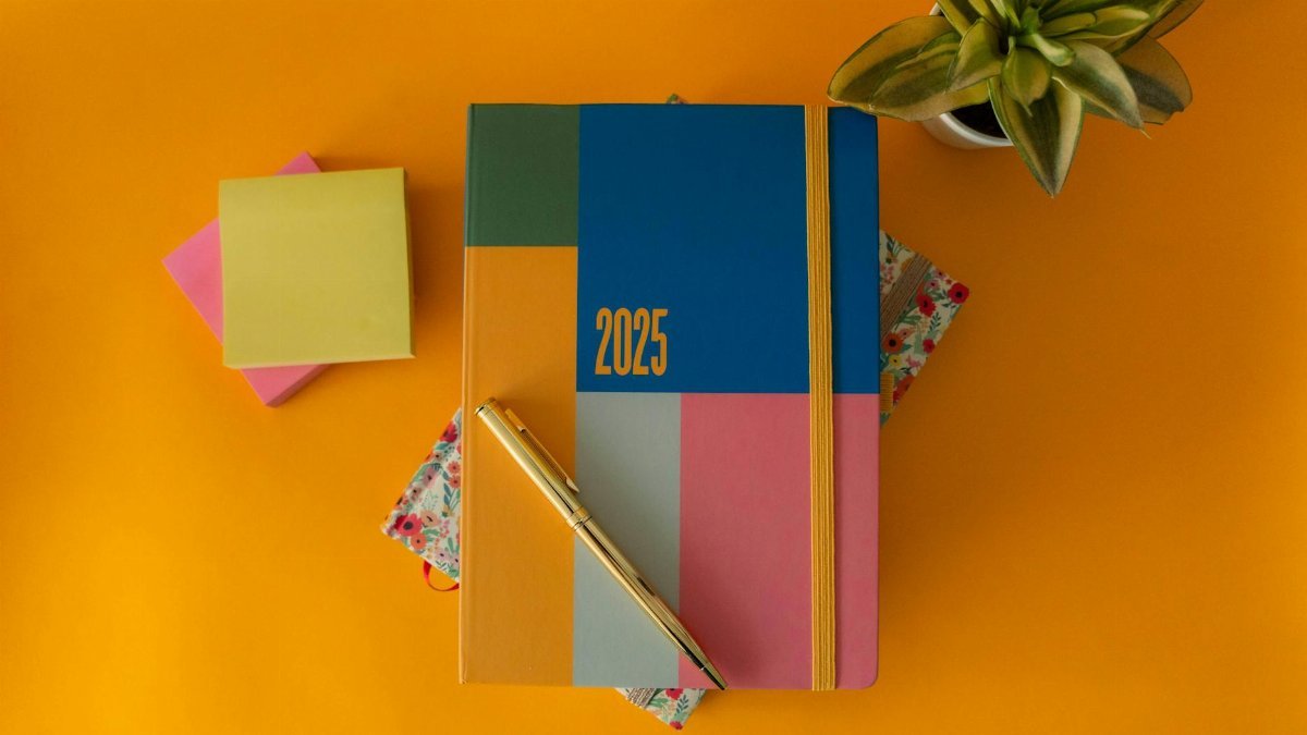 Flat lay of a vibrant 2025 planner with a pen and green plant on yellow background.