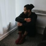 A contemplative man sits in a corner of a dimly lit bedroom, reflecting deep emotions.