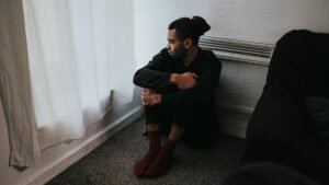 A contemplative man sits in a corner of a dimly lit bedroom, reflecting deep emotions.