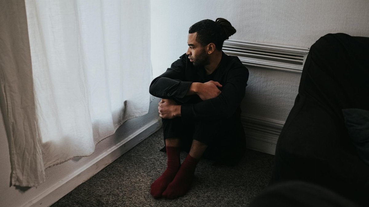 A contemplative man sits in a corner of a dimly lit bedroom, reflecting deep emotions.