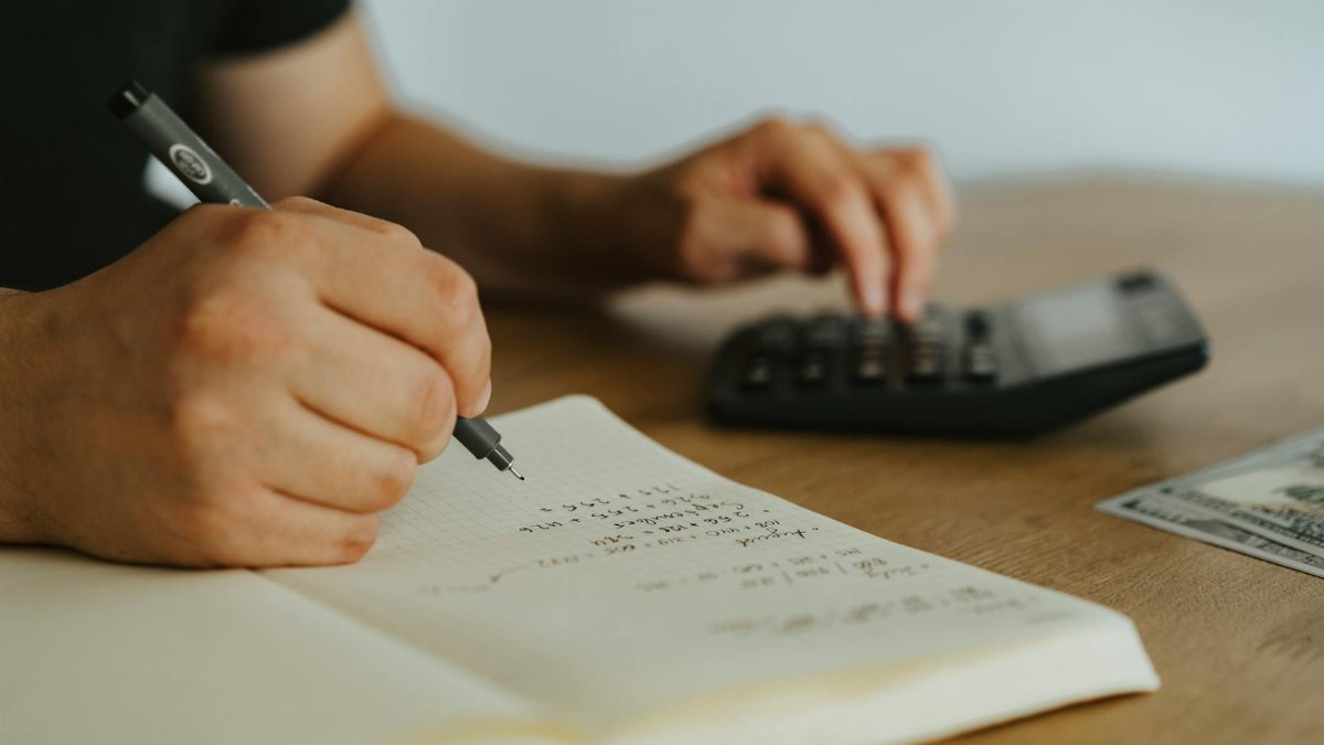 Close-up of hands writing calculations in a notebook with a calculator, focused on budgeting or financial work.