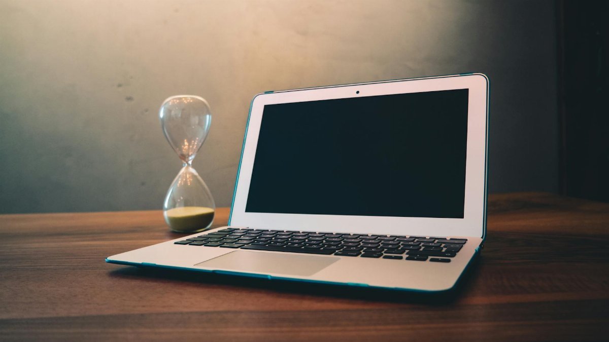Modern laptop on a wooden desk beside an hourglass, symbolizing time and technology intersection.