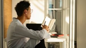 A young man sitting by a window in a sunlit home office, writing in a notebook.