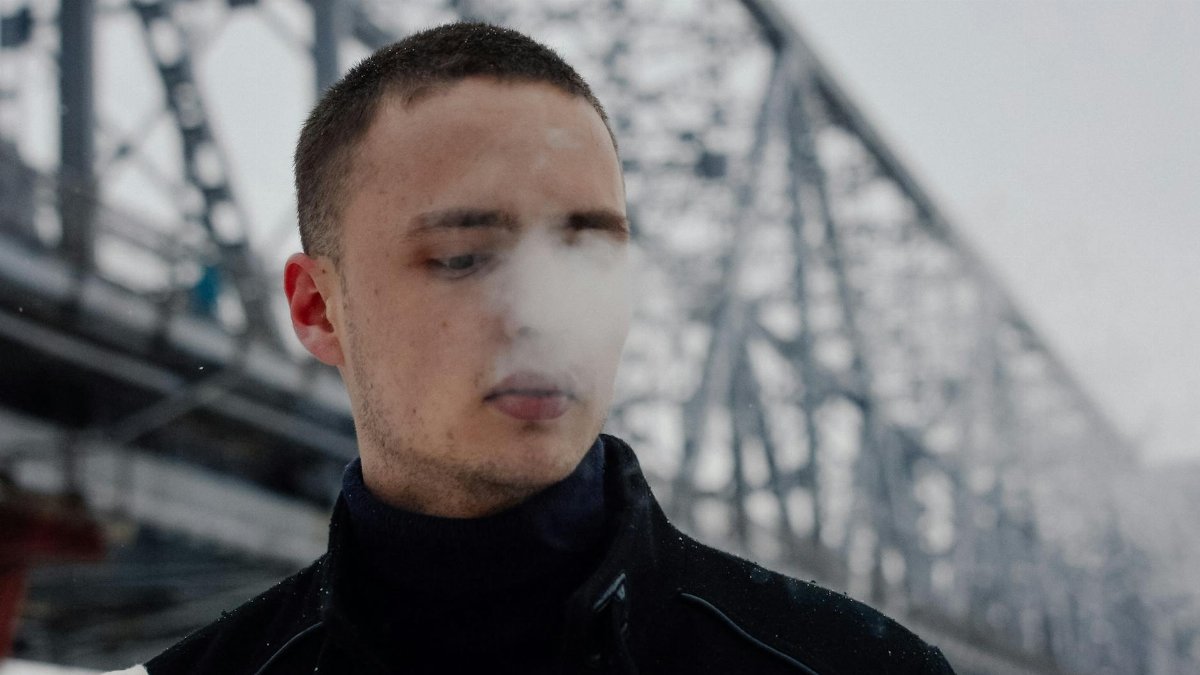 A young man exhales steam on a cold winter day, standing near a large iron bridge.