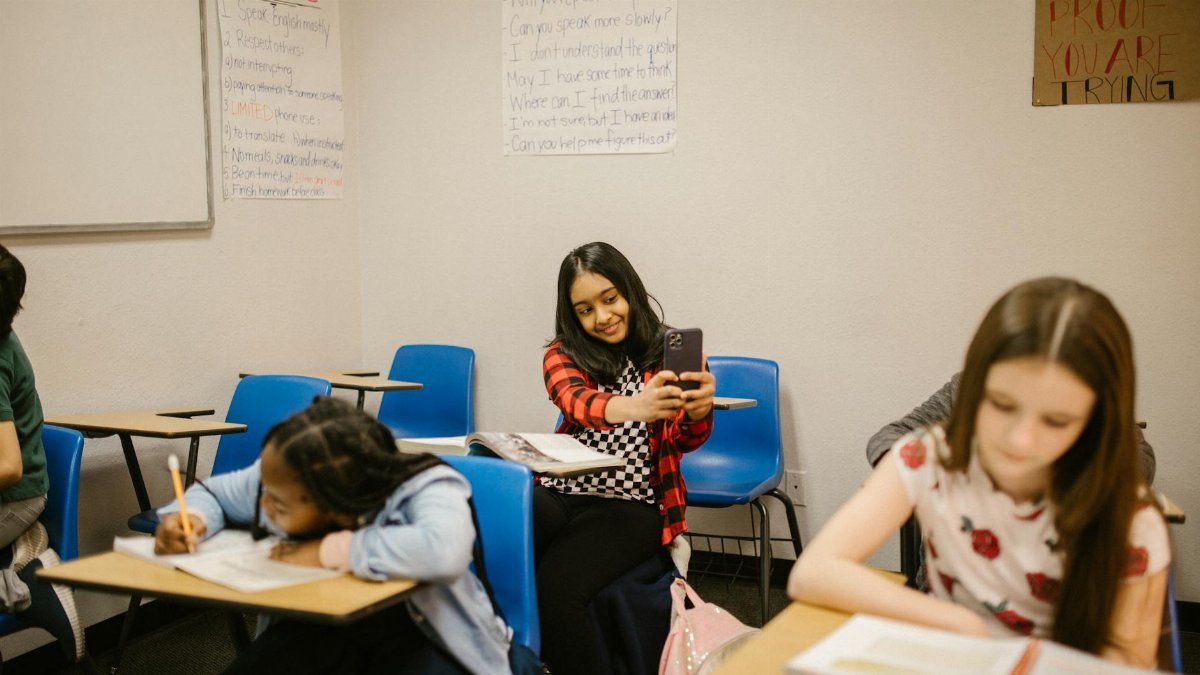 A diverse group of students in a classroom setting, one using a smartphone.