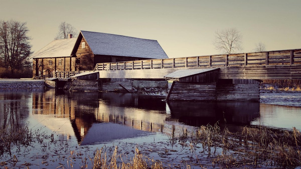 Rustic wooden house by a river with reflections and a wooden bridge in a serene autumn setting.