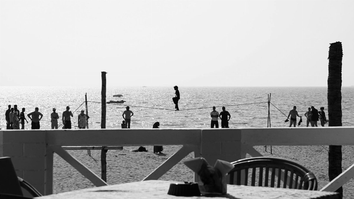 Monochrome image of a beach scene with a tightrope walker and onlookers by the sea.