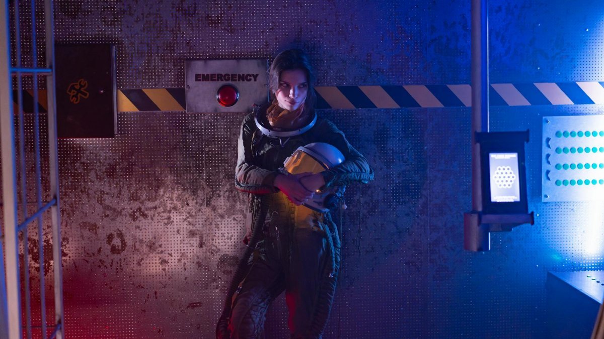 A woman in a spacesuit stands in a futuristic spaceship interior, illuminated by colorful neon lights.