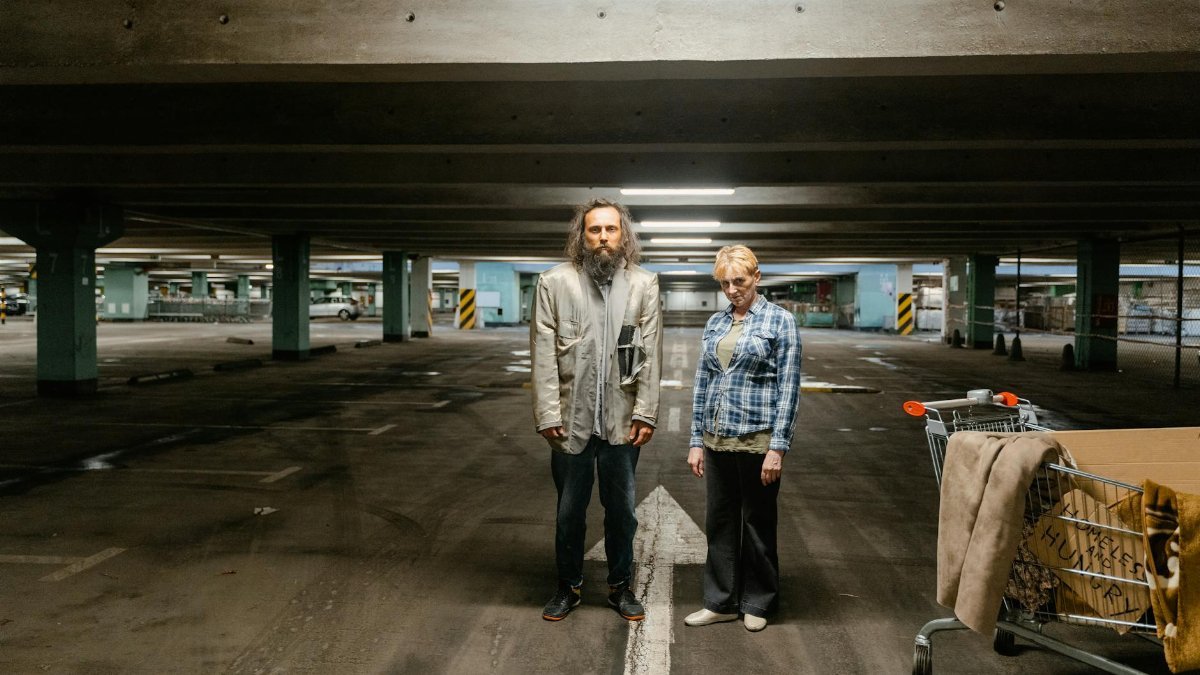 Elderly man and woman stand in a parking garage, symbolizing urban homelessness.