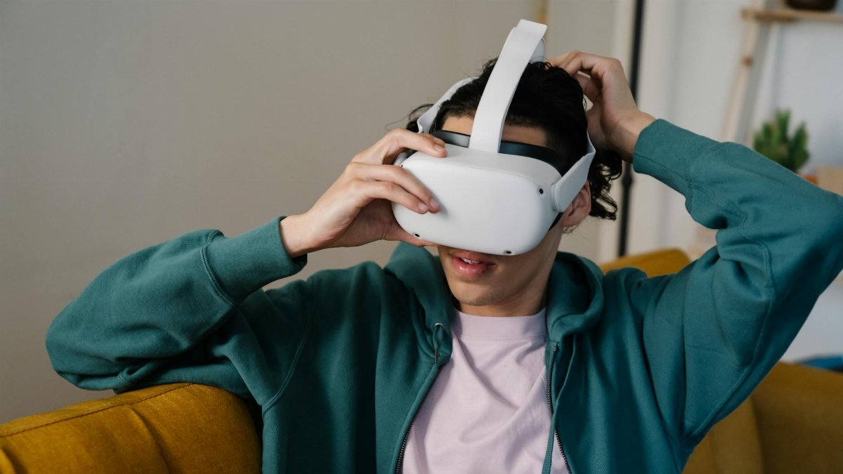 Male in hoodie putting on virtual reality headset while sitting on couch in light house