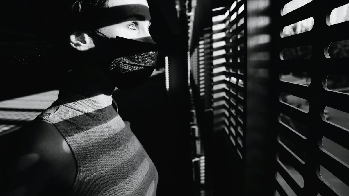 A woman wearing a mask looks out a window, casting dramatic shadows indoors.