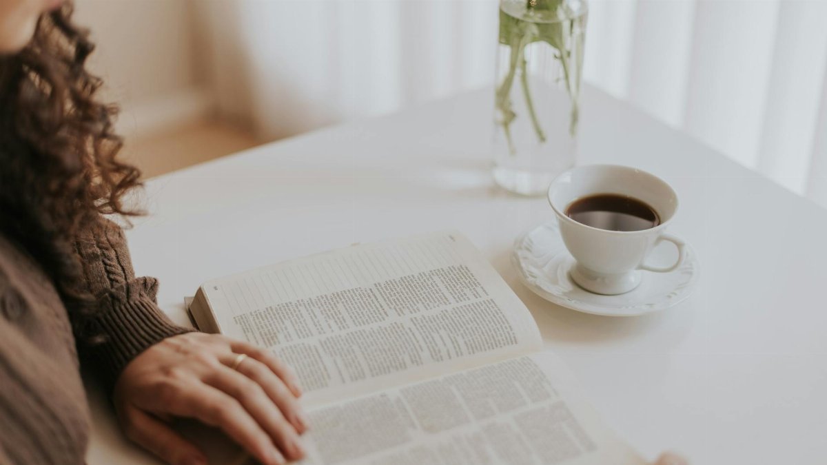 Woman reading a book at a white table with coffee and a vase, creating a cozy moment.