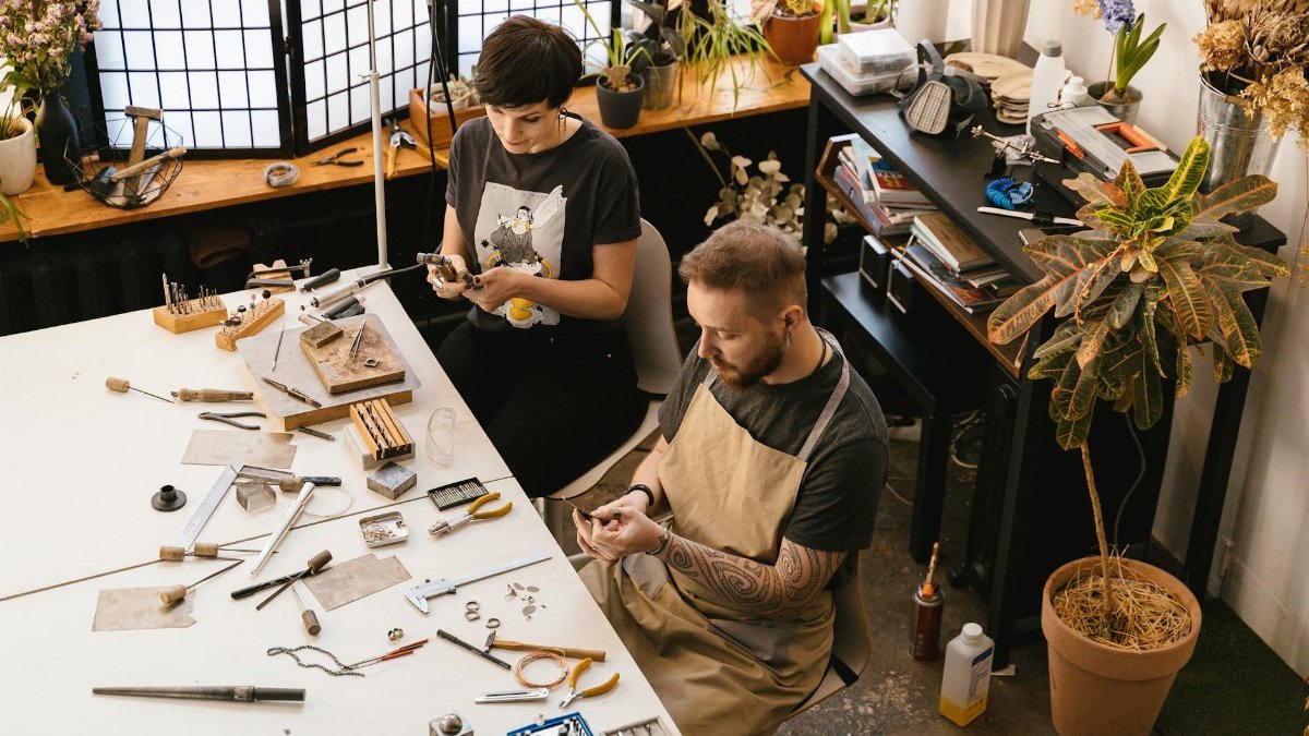 Two artisans working with tools and materials in a cozy, plant-filled workshop.